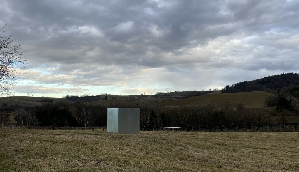 Eine Landschaftsaufnahme eines Feldes mit Bergen im Hintergrund. Die Forschungsstation, ein metallener Kubus, steht auf einer großen leeren Wiese.