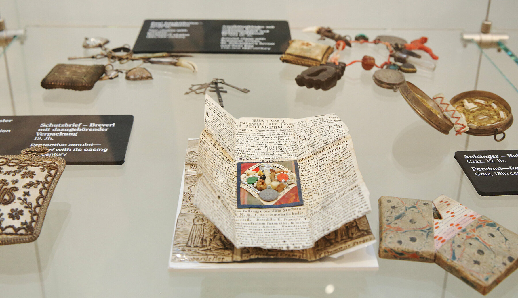 Ausstellungsansicht: historische Amulette und Schutzsegen auf Papier in einer Vitrine. Bis heute vertrauen Menschen auf einzelne Gegenstände oder Praktiken, die alle schädlichen Einflüsse abwehren sollen.