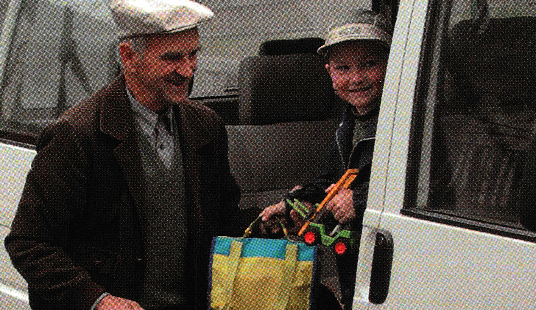 Ein Schulbusfahrer hilft einem Volksschüler mit bunter Schultasche aus einem Kleinbus, beide lächeln