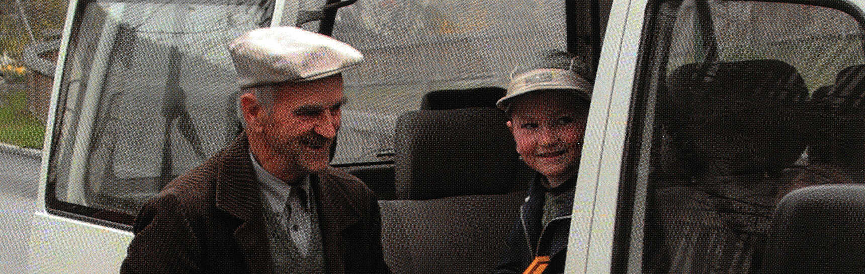 Ein Schulbusfahrer hilft einem Volksschüler mit bunter Schultasche aus einem Kleinbus, beide lächeln
