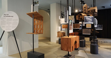 Ausstellungsansicht: Modelle aus Holz der verschiedenen Bauteile des Museumsareals, schwebend angebracht an Metallstangen.