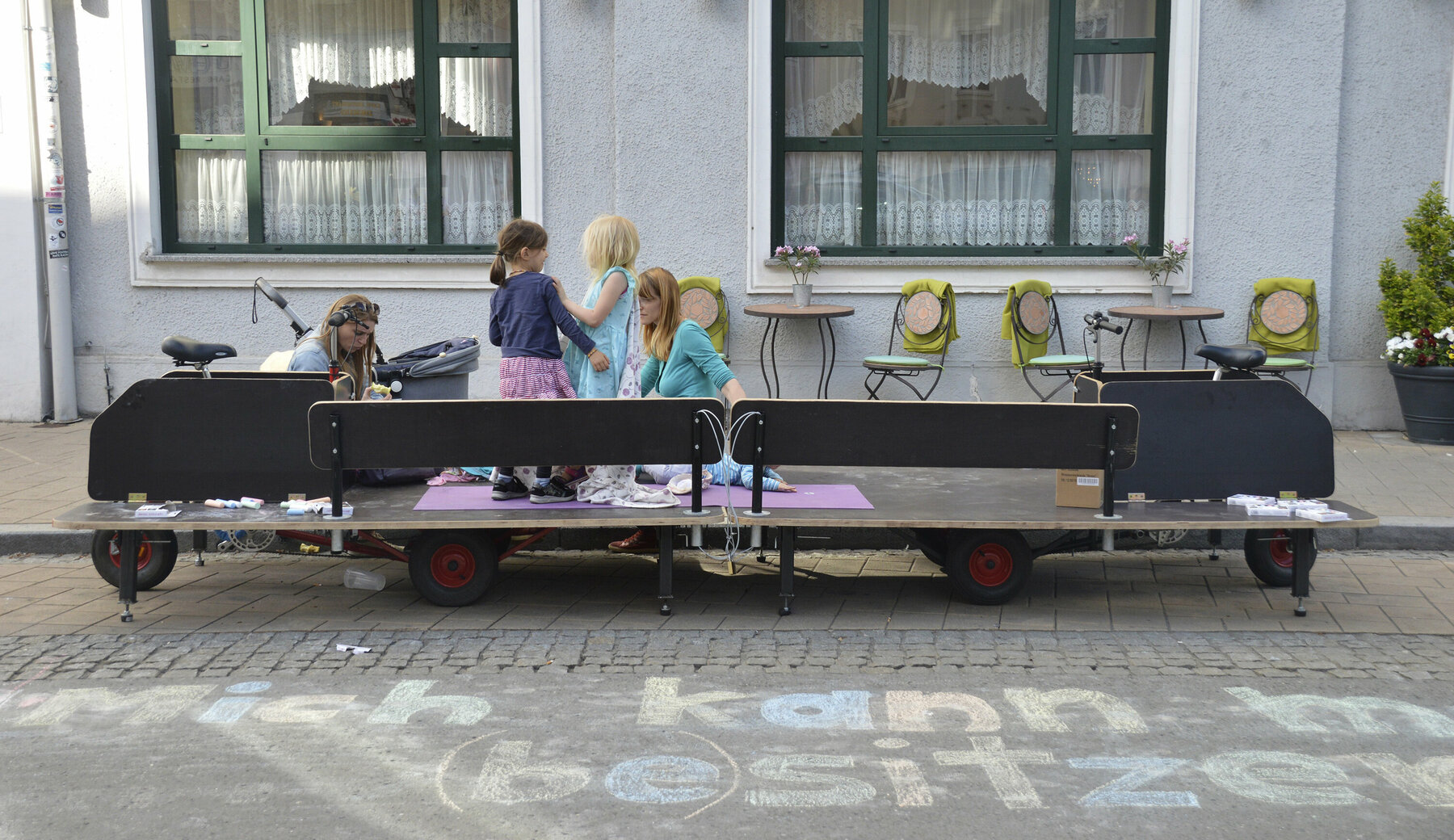 Eine große hölzerne Sitzmöglichkeit, die auch als Fahrrad funktioniert, parkt vor einem Haus in Graz. Zwei Frauen und ihre Kinder spielen darauf.