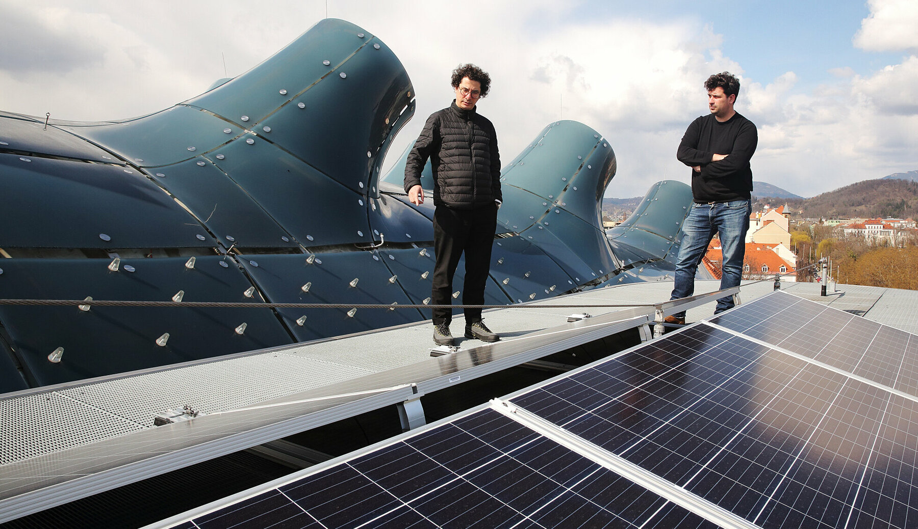 Die Medienkünstler Onur Sönmez und Jona Hoier stehen am Dach der Needle des Kunsthauses Graz und blicken auf Solarpaneele
