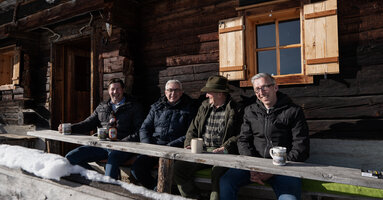 4 Herren sitzen auf einer verschneiten Bank vor einer traditionellen Holzhütte und unterhalten sich