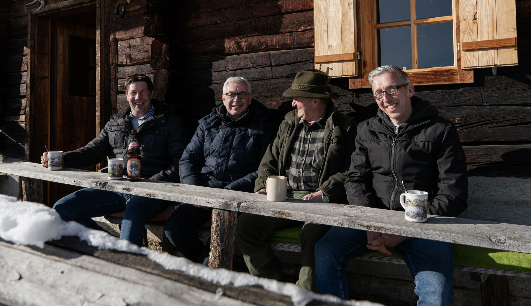 4 Herren sitzen auf einer verschneiten Bank vor einer traditionellen Holzhütte und unterhalten sich. 