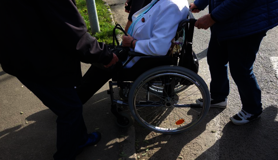 Einer Dame im Rollstuhl wird beim Spaziergang von zwei Menschen über eine Gehsteigkante geholfen.