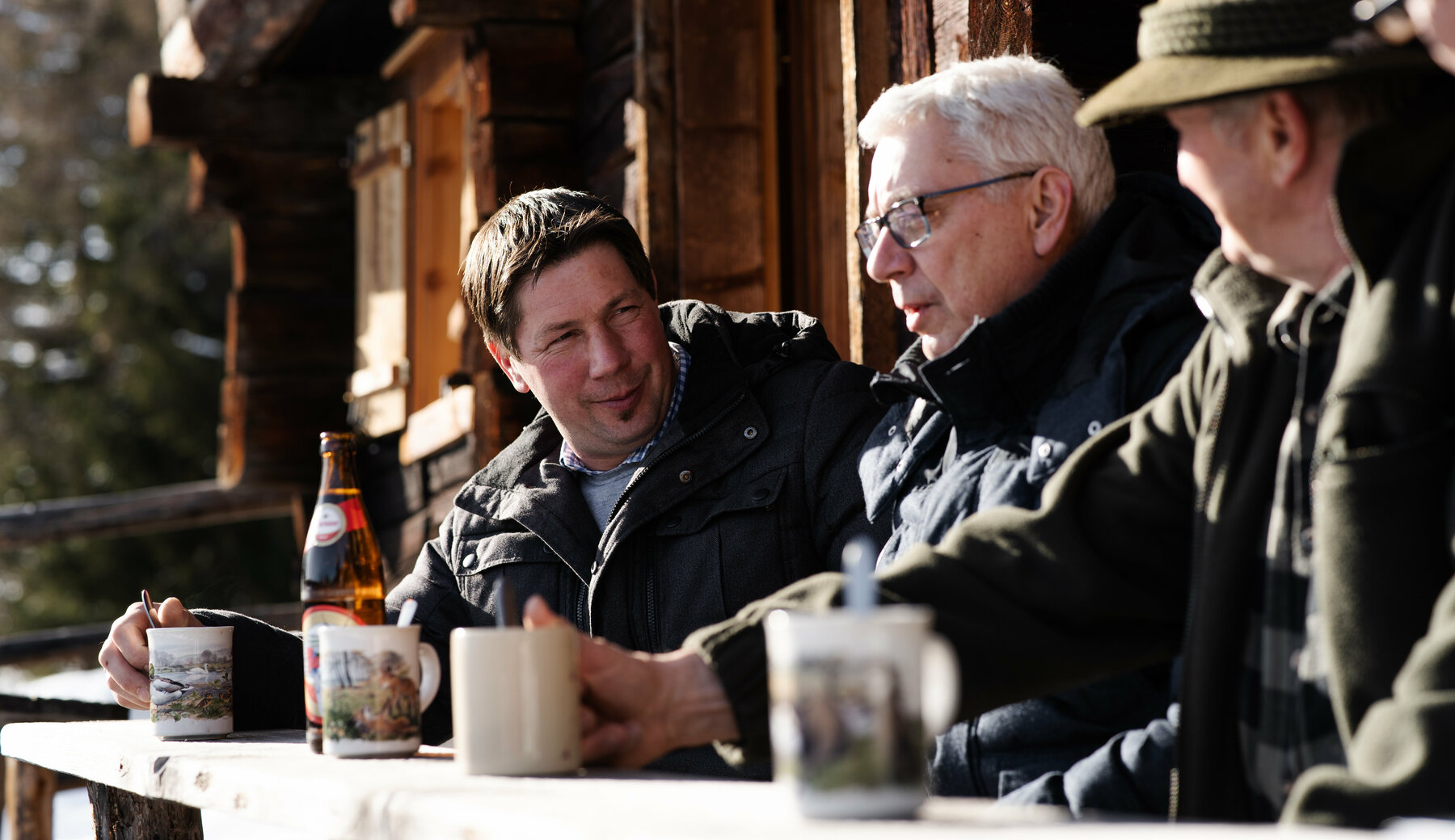 4 Herren sitzen auf einer verschneiten Bank vor einer traditionellen Holzhütte und unterhalten sich.